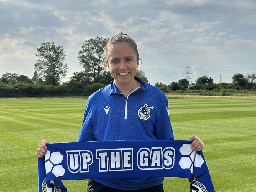 Chloe Bassett holding an 'Up The Gas' Scarf in front of her, on a football pitch.