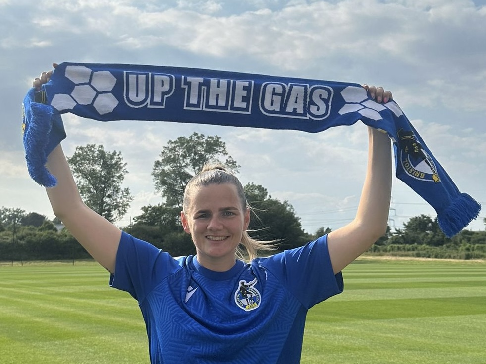 Rianne Bourne-Hallett holding an 'Up The Gas' Scarf above her head, on a football pitch.