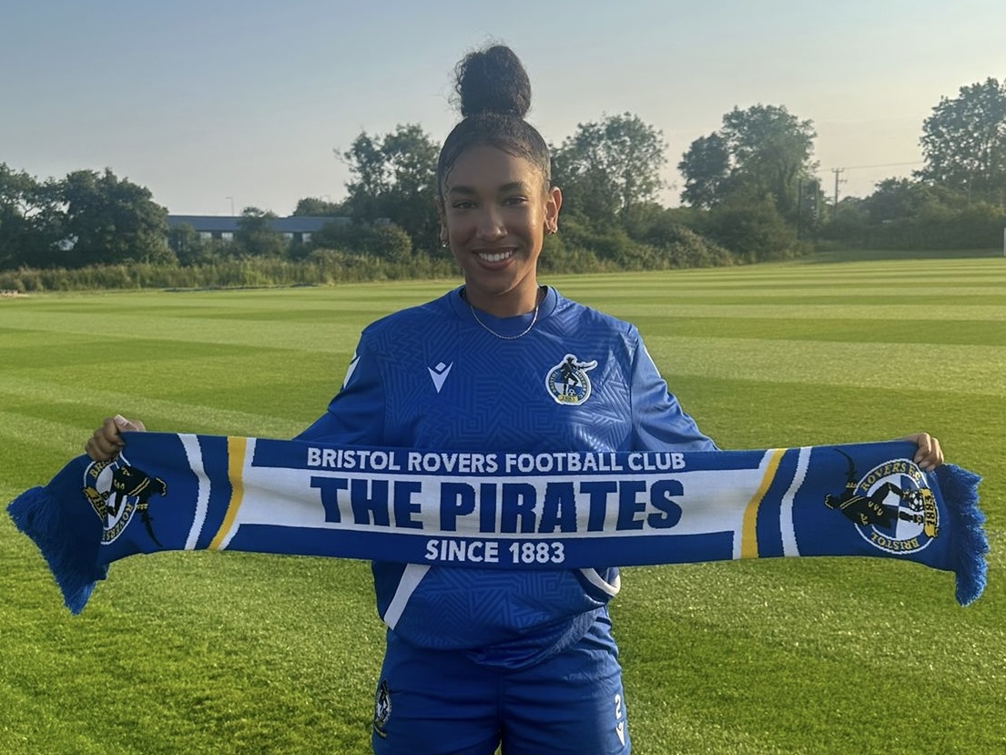 Layah Douglas holding an 'Up The Gas' Scarf above her head, on a football pitch.