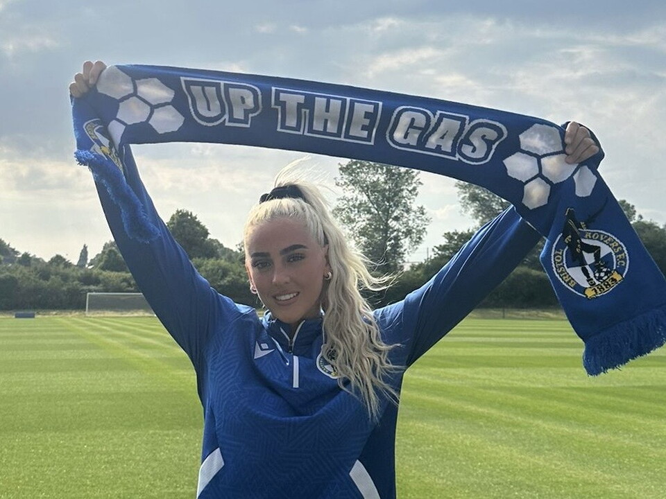 Daisy Ackerman holding an 'Up The Gas' Scarf above her head, on a football pitch.