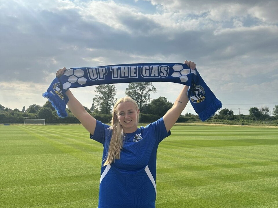 Meg Jarvis holding an 'Up The Gas' Scarf above her head, on a football pitch.